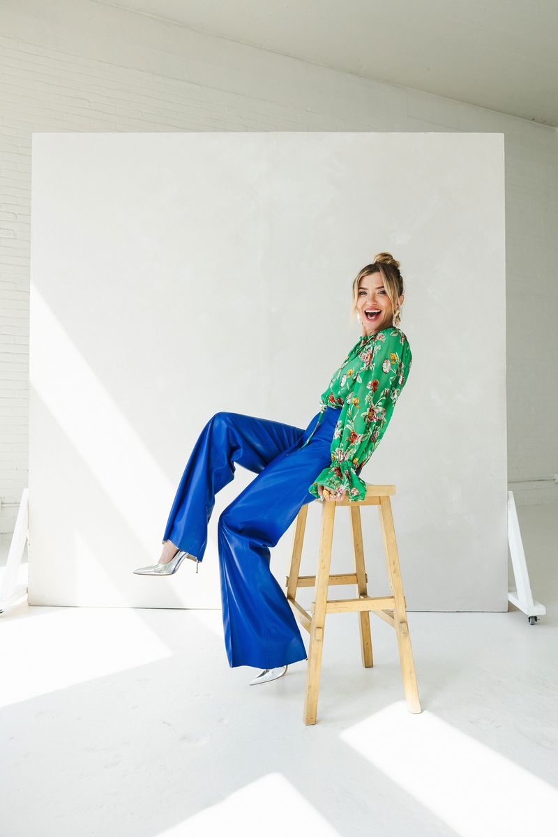 A woman sits joyfully on a wooden stool, wearing a vibrant green floral blouse, bold royal blue wide-leg trousers, and silver
